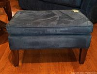 Front view of blue upholstered ottoman showing attached cushion and wood legs.