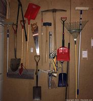 Full view of various yard and hand tools hung on a pegboard wall including shovels, saws, pickaxe, rake, squeegee and hand tools.