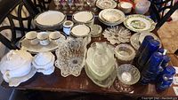 Full lot overview on dining table showing china, glassware, platters, soup tureen, various bowls, cups, and plates.