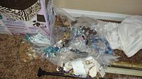 Photo showing multiple plastic bags filled with various styles of costume jewelry including metal, beaded, and colorful pieces on a floor corner.