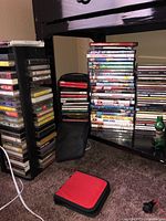 Stack of over 20 DVDs with colorful covers, a stack of CDs, and multiple cassette tapes stacked in racks along with a red and black carrying case on carpet.