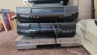 Stack of two black Panasonic VHS players on top of one silver Sony DVD/VHS player resting on carpet.