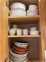 Cabinet shelves with white ceramic bowls, casseroles, and stack of various ceramic plates and bowls with coffee mugs on shelves.