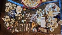 Overhead view showing assortment of miniature tea sets, spoons, a snow globe and other small collectibles in various materials and colors.