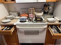 Kitchen countertop with stacked plates, casserole dishes, utensils in drawers, and various kitchenware items on stove and counter.
