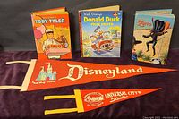 Photo of two red felt pennants, one Disneyland and one Universal City Studios, displayed alongside three vintage Disney hardback books including 'Toby Tyler', 'Donald Duck Prize Driver', and 'Zorro'.