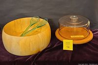 Wide view of glass domed cake/cheese plate with wooden base next to large wooden salad bowl with glass servers on dark fabric background.