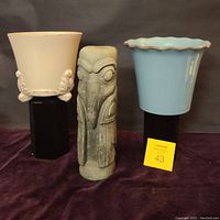 Full view of raven statue standing between two ceramic pots; cream footed pot on left, blue scalloped rim pot on right.