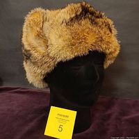Front and slightly right profile showing the plush natural brown and tan fur, rounded shape, and folded-up ear flaps.