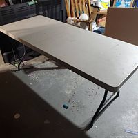 Photo of a rectangular collapsible table with a smooth beige plastic tabletop and black foldable metal legs in a folded position on a concrete floor.