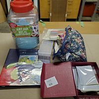 Overview of various crafting materials including a large plastic jar, floral fabric bag, small white storage containers, a scrapbook, and a red binder.