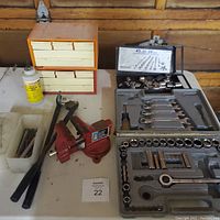 Photo of two socket sets, including ratchets, wrenches, and sockets, a red bench vice mounted on table, pipe bender tool, small plastic storage draws with hardware, and a bottle of chalk.