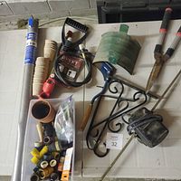 Photo of assorted items on a white table including hose connectors, hose fittings, a green snow shovel assist handle, a black wrought iron bracket, alumimium screening roll, BBQ rotisserie rods and motor, and gardening accessories.