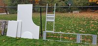 Disassembled parts of the Ikea Gallant desk system laid on grass near fence, showing curved and rectangular white desktop panels and metal frame components including legs and frame sections.