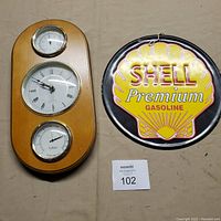 Front view of the wooden wall mount with clock, thermometer, and hygrometer next to the Shell Premium Gasoline metal sign.