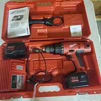 Milwaukee cordless drill kit inside red hard plastic case showing drill, battery, charger, drill bit holder and manual.