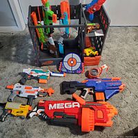 Full view of eleven Nerf blasters laid on carpet in front of black storage rack and target game.