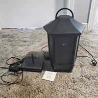 Photo showing the black hexagonal lantern-style wireless speaker, black wireless transmitter with antenna, power adapter, and connectors on carpeted floor.
