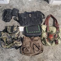 Five used purses of varying styles and colors displayed on carpet, including black leather with skull emblem, camouflage bag, brown bag with multiple pockets, black studded bag, and canvas tote with eagle design.