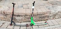 Two kick scooters resting side by side on pavement in front of a low stone wall, showing overall condition and size difference.