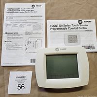 Front view of Trane touchscreen programmable thermostat with screen off, accompanied by instruction manual and owner's guide.