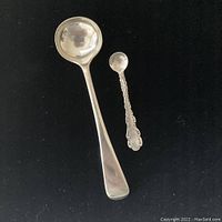 Photo showing two silver spoons on black background, one large ladle spoon and one smaller ornate spoon.