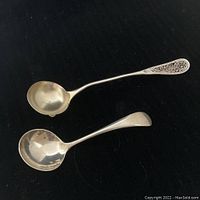 Two sterling silver ladles, one with filigree handle detail and one with plain handle, placed on dark background showing size and shape.