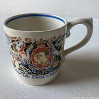 Side view of the mug showing profile portraits of King George VI and Queen Elizabeth, along with other colorful royal commemorative imagery on cream background.