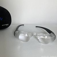 Front view of MaxTV vision glasses showing the clear lenses and black arms alongside the branded carrying case.