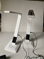 Pair of white adjustable light temperature table lamps with hinged arms, featuring rectangular bases and slender arms with LED light strips at the top, photographed against a plain background.