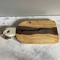 Wooden charcuterie board with white marble handle and metal cheese tool on top, showing overall look and size.