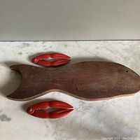 Photo showing the full lot: one wooden fish-shaped charcuterie board and two red lobster claw crackers on a white marble surface.