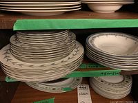 Shelves holding two stacks of Aynsley china plates and bowls with two flower and leaf patterns, one with blue leaf border and silver rim, other with green leaf floral rim.
