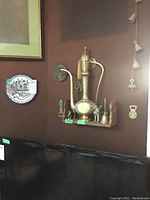 Wall-mounted wooden shelf holding a large detailed brass ewer, brass figurines, small bell, and plate mounted nearby. Hanging brass chimes visible on adjacent wall.