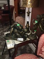 Two bronze-like sculptures on marble top table, one sculpture with two intertwined male figures, other is standing male figure in classical pose; bust sculpture in background.