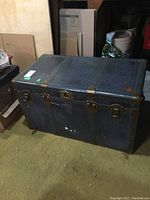 Closed metal chest trunk showing riveted dark metal exterior, latch closures, and side handles.