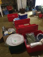 Wide angle photo showing two large coolers (one blue, one white), several red plastic bins containing disposable platters, napkins, and table cloths in garage space.