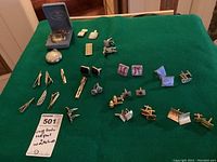 Wide shot showing all cuff links, tie pins, and two pocket watches on a green felt surface with seller note indicating items included.
