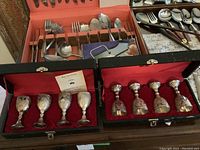 Two black lined cases with eight silver plate British-style goblets arranged inside, five in one case, three in another, with intricate relief designs.