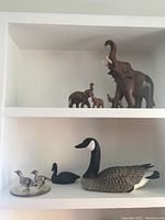 Wood carved elephant figurines displayed on a white shelf, one large elephant with trunk raised and three smaller elephants beside it. On the lower shelf, ceramic bird figurines including two loons and two ducks.