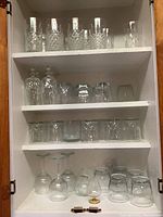 Photo of the glass cabinet with multiple shelves holding various crystal and glass drinkware including tumblers, goblets, and tall boys featuring crystal cuts.