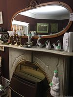 Wide photo of fireplace mantel showing multiple Lladro figurines, brass-look candelabra, oval ornate mirror and Buddha statue on pedestal below