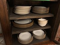 View of stacked Mikasa Lorenzo A1-128 white dishes with gold rims and Noritake Crestwood Cobalt Gold 4171 dishes on wooden shelf.