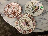 Top view of three decorative dinner plates showing floral and bird motifs with scalloped edges on marble table.