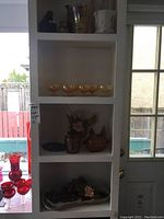 Four shelves displaying amber aperitif glasses, various glass and decorative vases, a candle holder figurine, match holder, and an oval tray with artificial grapes and flower.