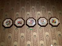 Five floral collector plates arranged horizontally on a patterned wallpaper, showcasing colorful flower artwork on each plate, framed in dark octagonal wood frames.