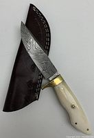 Full view of the Damascus steel knife resting on leather sheath, showing blade with Damascus pattern, white handle with rivets, and brass bolster.
