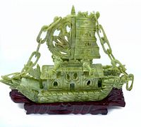 Front view of green carved jade sailing ship on reddish-brown carved wooden stand.