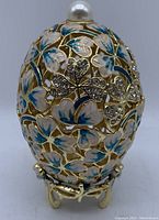 Front view of gold-tone egg trinket box with turquoise and white enameled butterflies and clover leaves accented with rhinestones, pearl-like finial on top.