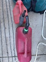 Two red gas cans with black spouts, one with green tape on spout, placed on concrete